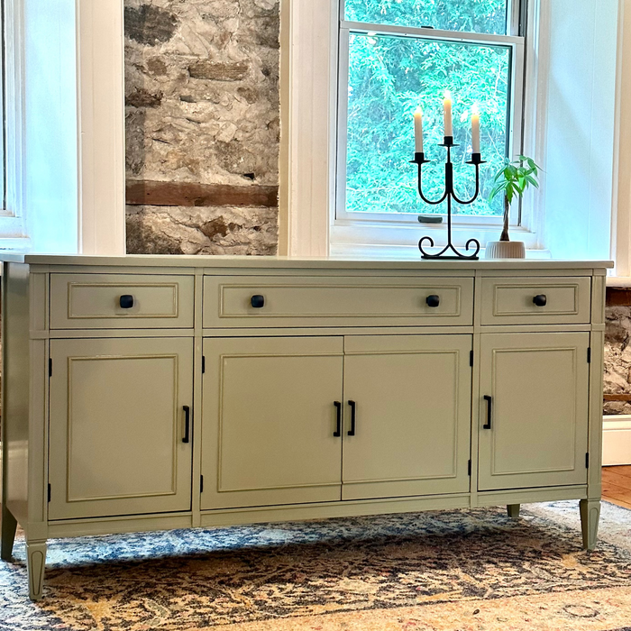 Green sideboard with candle holder and plant in a room with stone wall and window.