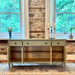 Light green wooden console table with open doors against a stone wall and large windows.